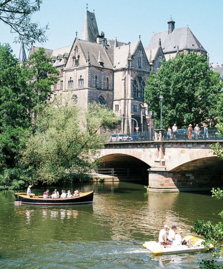 Neugotisches Gebäude über der Lahn, davor eine Steinbrücke, ein Ausflugsboot und ein Tretboot auf dem Wasser